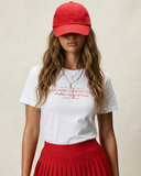 Woman wearing a red cap, white t-shirt with text, and red pleated skirt on a beige background
